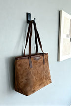 Brown tote bag hanging on a wall hook against a light gray wall.